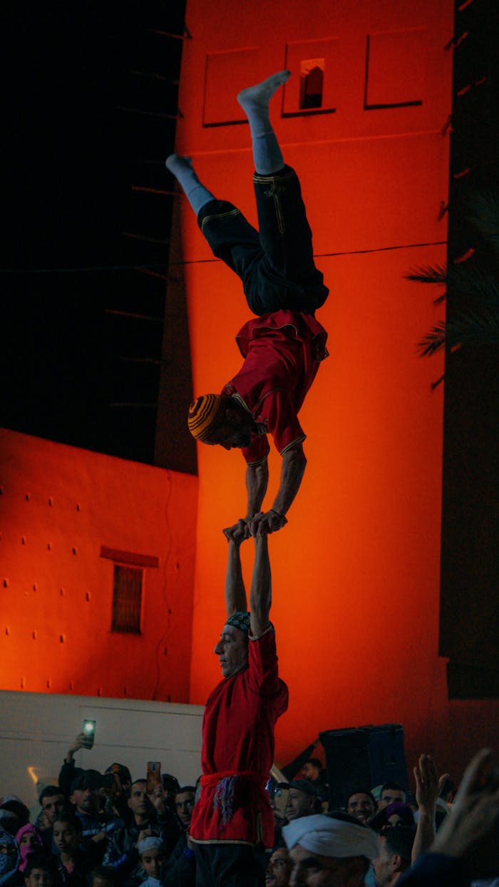 service-03 Two acrobats perform a balancing act in front of an illuminated building at night, captivating the audience.